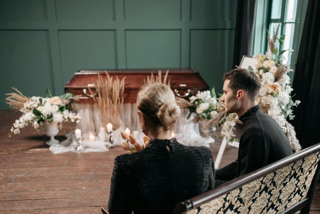 A funeral service with a wooden coffin, adorned with flowers and candles, attended by two mourners.