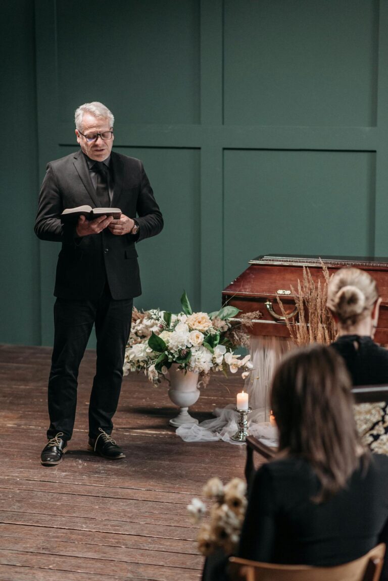 A solemn indoor funeral ceremony with pastor, family, and floral arrangements.